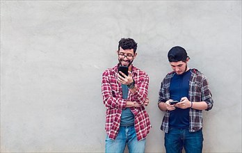 Two smiling friends leaning on a wall checking their cell phones