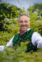 Laughing middle-aged man in Bavarian traditional costume standing by a hedge