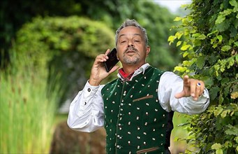 Man with telephone in Bavarian traditional costume at a hedge