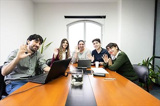 Group of employees sitting in office meeting room looking at camera and waving. Online meeting concept