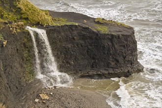 Waterfall flowing over hard shale to beach on rough sea coast
