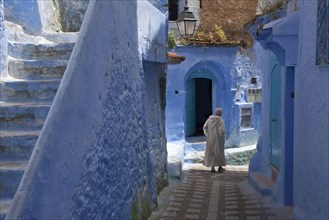Man with djellaba walking next to blue buildings and staircase in an alley of the city