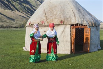 Two Kazakh woman in traditional dress in front of a yurt greeting guests with sweets
