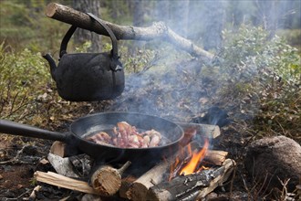 Blackened tin kettle boiling water and pan cooking bacon over flames from campfire
