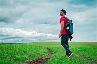 Side view of backpacking man on a beautiful road in the countryside. Young explorer man walking in a green field