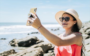 Girl in hat on the beach taking a selfie. Young woman on vacation taking photos on the beach