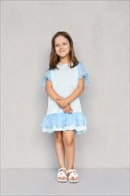 Cute little girl in blue dress and white sandals posing with hands clasped down near the gray wall