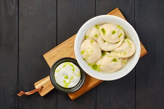 Overhead view of a bowl with beef pelmeni with sour cream on a wooden serving board