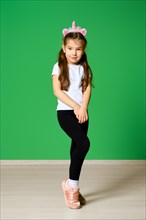Funny little kid with unicorn hair hoop on head and in white t-shirt and black leggings posing over green background
