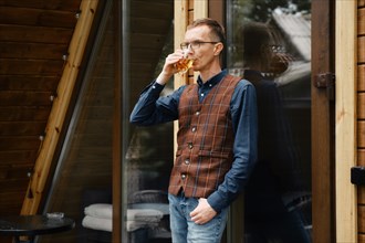 Stylish middle-aged man stands on the terrace of a forest cabin leaning against glass door and drinks whisky