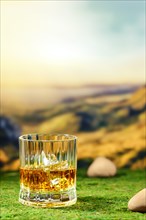 Facetted glass of smoky whiskey placed on green moss with defocused mountain landscape on background