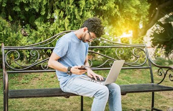 Young man in a park with laptop and cell phone. Freelancer man sitting in a park using laptop and cellphone. Man in a park working online with laptop. Relaxed man working with laptop outdoor