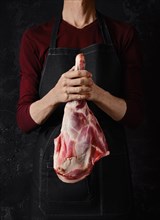 Butcher holds raw lamb leg in hands over dark background