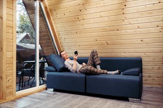 Man rests on sofa with smartphone in his forest cabin