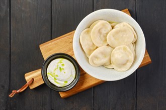 Overhead view of a bowl of pelmeni with curd with sour cream on a wooden serving board