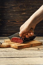 Hand with knife cutting smoked dried beef meat on slices on wooden cutting board