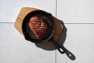 Top view of grilled beef steak in cast iron skillet on ceramic tile background with harsh shadow