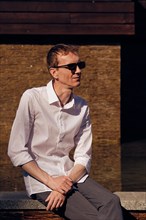 Middle age man in white shirt and sunglasses sitting in shade in hot sunny day