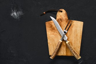 Overhead view of wooden cutting board with steak knife and fork on black shabby background