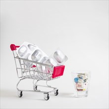 Many pills small glass with shopping cart with silver blister pills white background