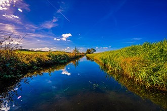 A small irrigation canal between two fields with reflection of clouds and the blue sky in autumn
