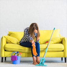Frustrated young woman sitting sofa with cleaning equipments