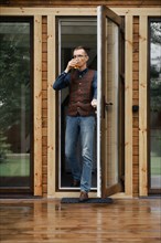 Stylish middle age man comes out on the terrace of a small wooden house with a glass in hand