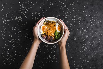 Elevated view hands holding bowls ramen noodles with eggs salad spread with rice grains black background
