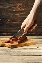 Hand with knife cutting smoked dried beef and deer sausage on slices on wooden cutting board