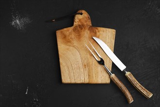 Knife and fork for steak on empty wooden cutting board over black background