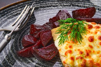Close up view fried cod fillet with melted cheese topping and roasted beetroot slices on dark wooden table