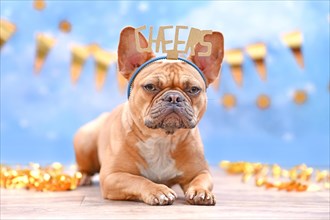 French Bulldog with birthday part headband with words Cheers in front of blurry blue background with garlands and party streamers
