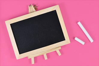 Wooden empty black chalkboard next to pieces of white chalk on pink background