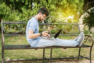 Freelancer man sitting in a park using laptop and cellphone. Man in a park working online with laptop. Relaxed man working with laptop outdoor