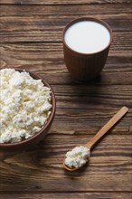 Fresh cottage cheese in clay bowl with wooden spoon with a glass of milk on rustic wooden background