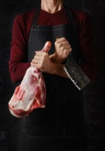 Butcher holds raw lamb leg in one hand and cleaver in another over dark background