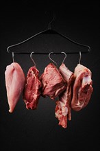 Different cuts of raw meat on hook on hanger over black background