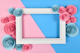 Cute flat lay with empty white picture frame surrounded by romantic paper craft roses on pastel blue and pink background