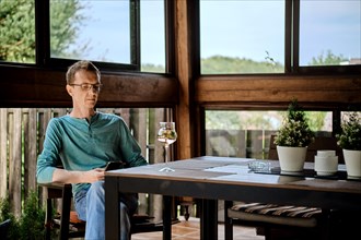 Middle aged man sits on the veranda of country house with a glass of wine and uses smartphone