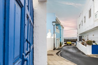 Yellow 4-wheel drive car on narrow street with white and blue buildings in Corralejo