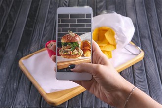 Making photo with phone of bacon burger with nachos on wooden plate. Food blogging