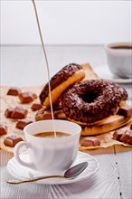 Pouring coffee in cup on the table with chocolate donuts on bright wooden background