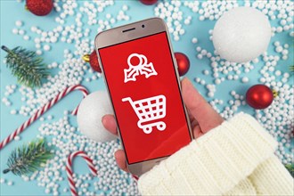 Concept for Christmas seasonal online gift shopping with hand holding cell phone with white shopping cart sign on red background in front of seasonal decoration