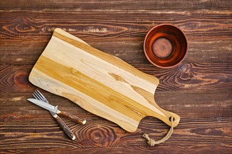 Top view of empty cutting wooden board with clay bowl and fork with knife on the table