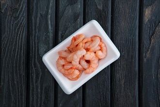 Small boiled peeled shrimps in small plate on dark wooden background. Top view