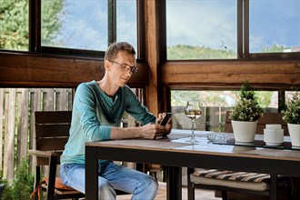 Smiling middle aged man drinks wine sitting on the veranda of his country house and using smartphone