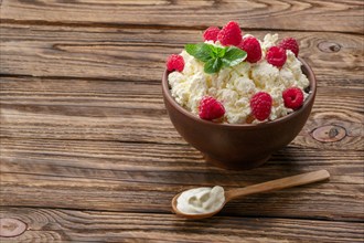 Wooden spoon with sour cream near clay plate with fresh cottage cheese with raspberries and mint