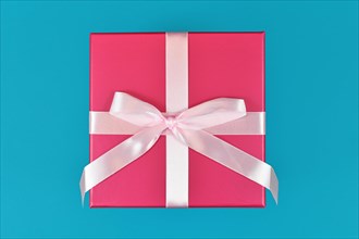 Single square shaped pink gift box with tied ribbon bow on teal blue background
