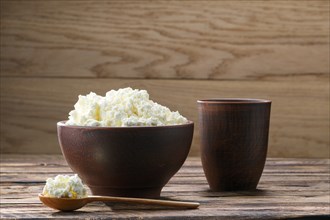 Fresh cottage cheese in clay bowl with wooden spoon with a glass of milk on rustic wooden background