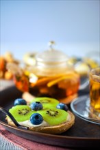 Selective focus photo of cottage cheese on fresh bun with kiwi and blueberry and tea on background